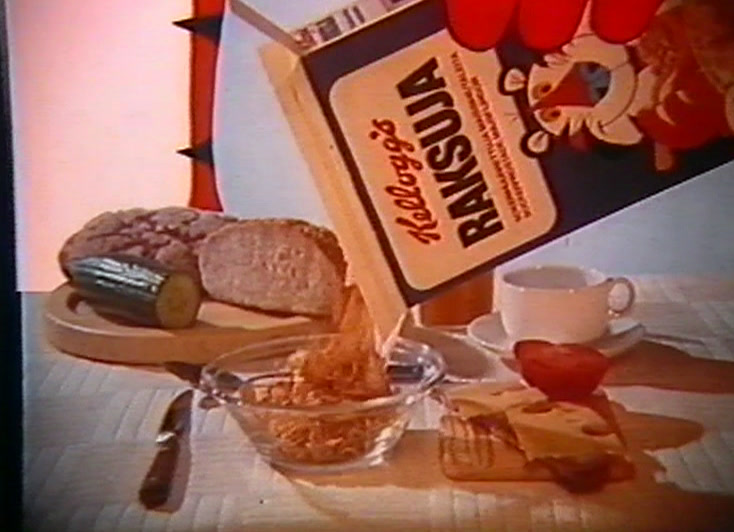 Aamiaispöytä, jossa on leipää ja voita, ja Kellogg’s Raksuja -paketti kallistettuna kameraa kohti.