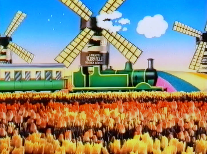 Two windmills stand behind the green train above the wide flower field.