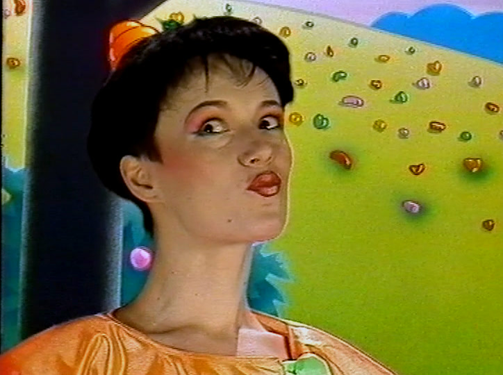 A woman with a black bob in an orange top looks up at a yellow-green candy hill.