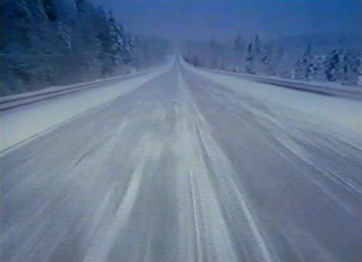 Luminen tie kulkee suoraan talvisen metsän halki, kuvattuna liikkuvasta autosta.