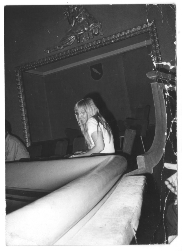 Marjut Rimminen, young, with long fair hair and glasses, turns in a theatre balcony at Annecy, smiling, the ornate proscenium above her.