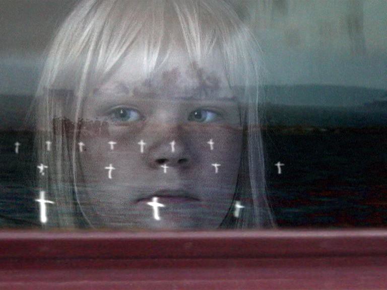 Still from Learned by Heart, a child's face behind glass, rows of small white crosses reflected across it
