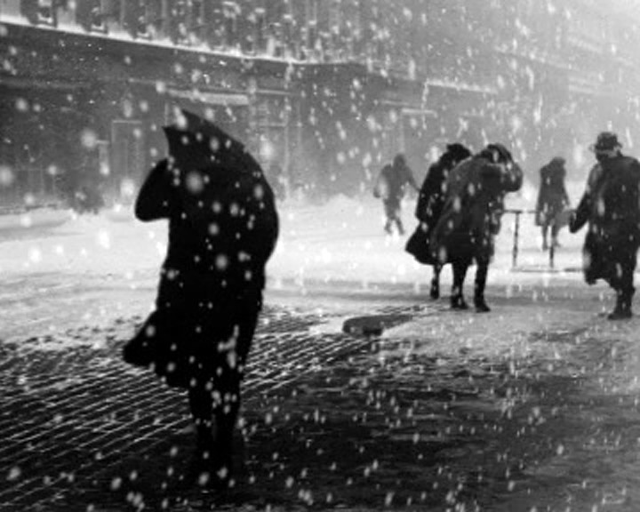 Still from Learned by Heart, people in coats and hats pushing through heavy snowfall on a city street, one carrying an umbrella