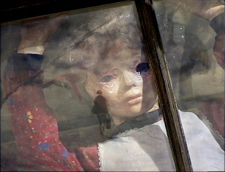 Still from Many Happy Returns, a puppet girl in a red flowered dress pressed against a window, a walking figure reflected in the glass