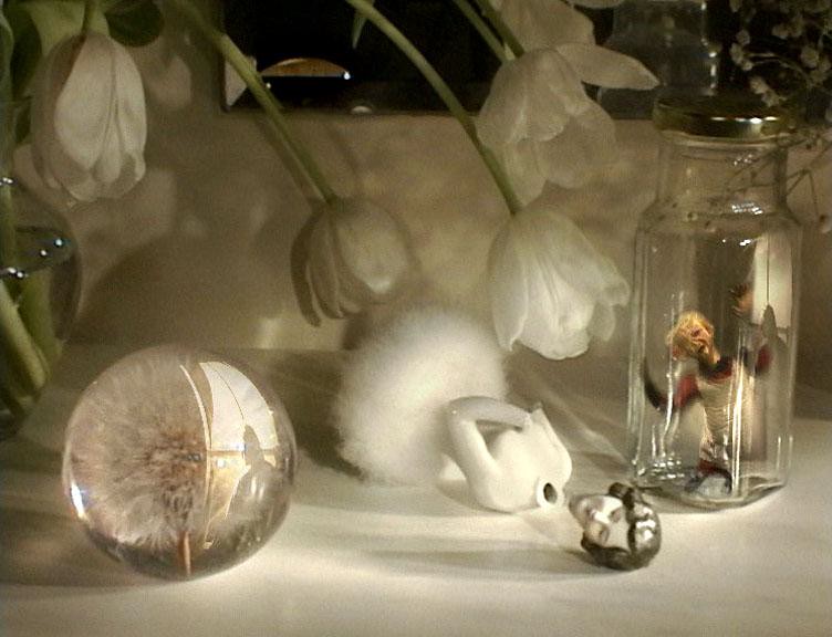 Still from Many Happy Returns, white tulips, a dandelion clock in a glass sphere, a china rabbit and a tiny figure inside a glass jar