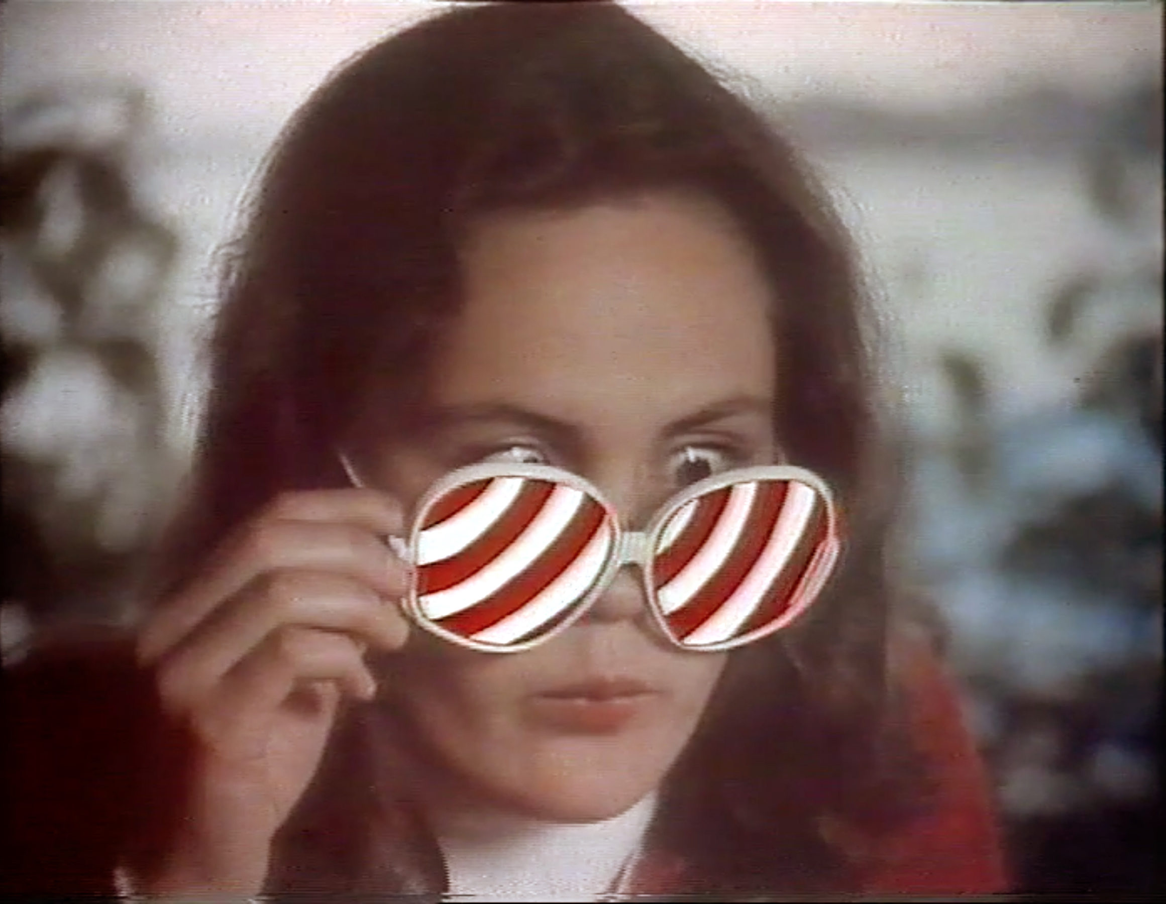 Marianne still, a woman with long dark hair holds a pair of large round white-rimmed sunglasses up to her face and both lenses are filled with red and white candy stripes, her eyes looking out above the frames, blurred greenery behind