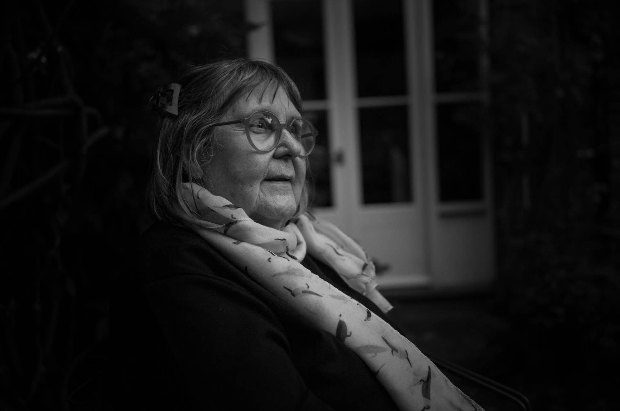 Marjut Rimminen istuu lasisten pariovien ääressä iltahämärässä, pyöreät silmälasit nenällään ja kuviollinen huivi kaulassaan
