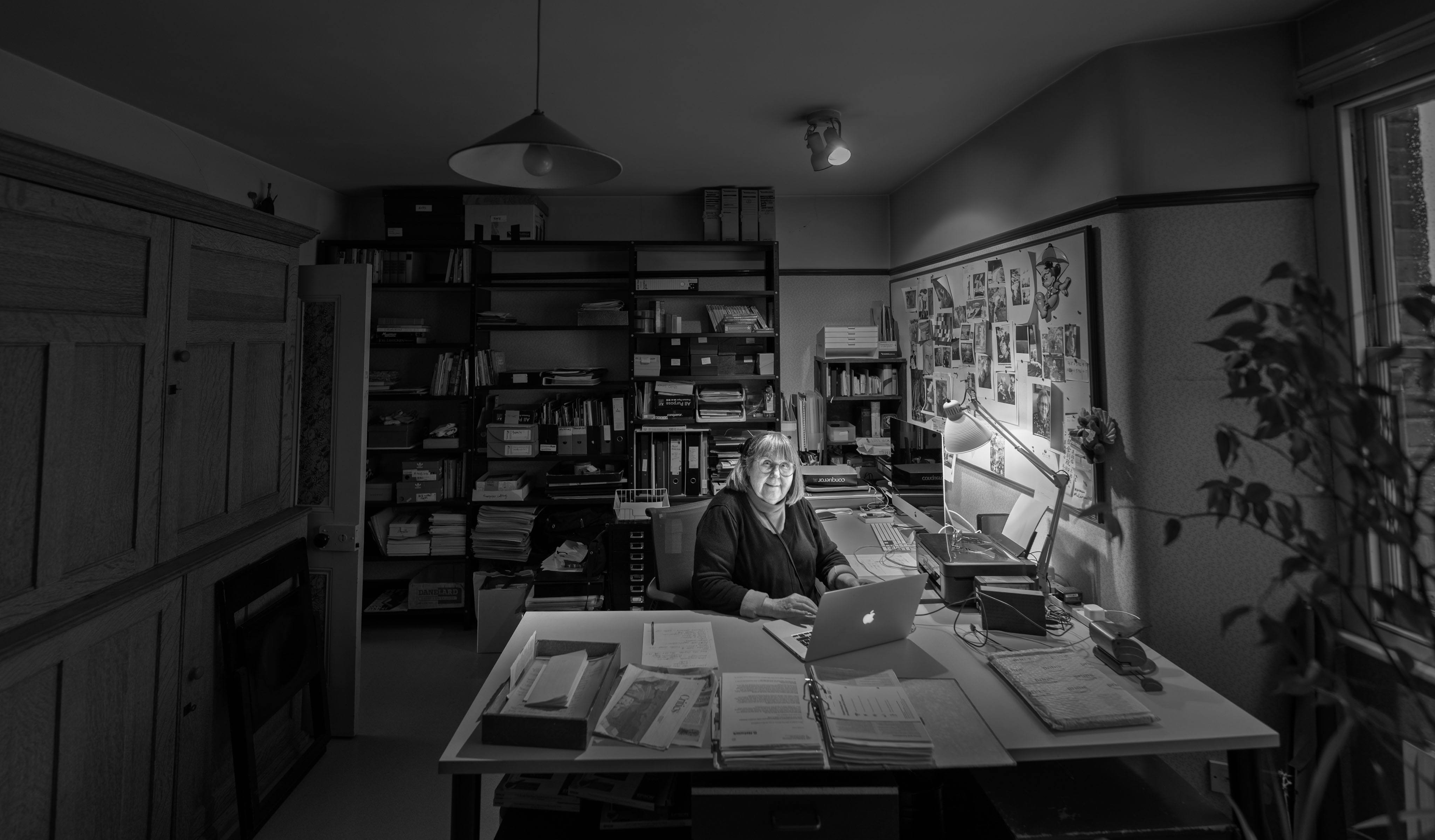 Marjut Rimminen työpöytänsä ääressä Lontoon studiossaan, ympärillään arkistohyllyjä ja lehtileikkeiden peittämä korkkitaulu